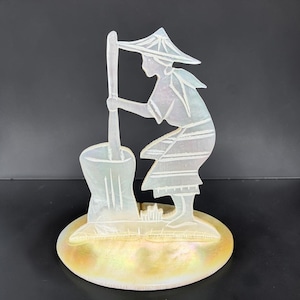 May include: A carved mother-of-pearl figurine depicting a person in a conical hat using a mortar and pestle. The figurine is mounted on a pearlescent oval base. The figure is light in color and the base has a golden hue.