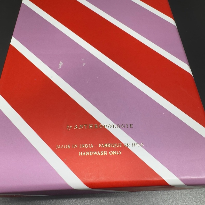 May include: A box with a diagonal striped pattern in red, white, and lavender. The text "ANTHROPOLOGIE" is printed in gold. The text "MADE IN INDIA - FABRIQU&Eacute; EN INDE HANDWASH ONLY" is also printed in gold.