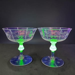 May include: Two vintage champagne coupe glasses with a green stem and base. The clear glass has a floral and grid pattern. The glasses have a slight iridescent sheen, with green and purple hues. The stem and base glow green.
