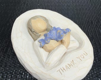 May include: A carved, off-white figurine of a person holding blue flowers. The figure is on an oval base with the words "THANK YOU" carved into the base.
