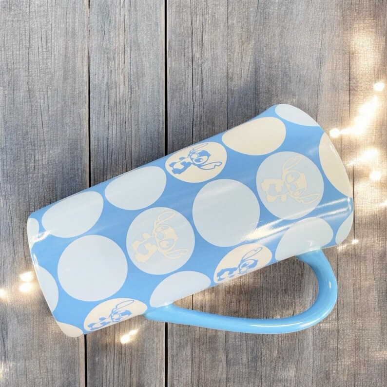 May include: A blue and white polka dot mug with a light blue handle. The mug features a repeating pattern of white circles with a blue cartoon character inside each circle.