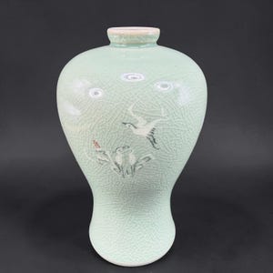 May include: A celadon-colored ceramic vase with a crackle glaze. The vase features a painted design of birds and foliage. The vase has a rounded body and a narrow neck. The vase is approximately 8 inches tall.