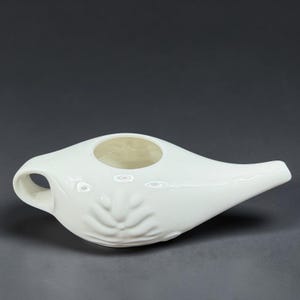 May include: A white ceramic neti pot with a glossy finish, featuring a rounded body, a tapered spout, and a small handle. The surface is decorated with a subtle, embossed floral or leaf-like pattern on the side, and it has a circular opening.