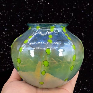 May include: A small, round, iridescent glass vase with a light blue and green color gradient. The vase is decorated with green polka dots.