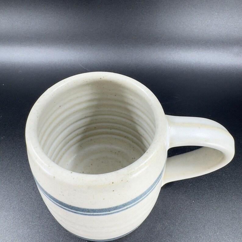 May include: Cream-colored ceramic mug with a blue stripe around the middle. The mug has a rounded shape with a handle and a textured surface. The interior of the mug is also cream-colored.