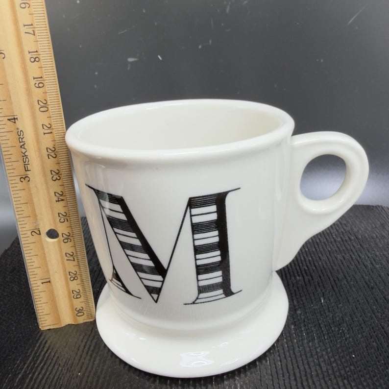 May include: White ceramic mug with a black and white striped letter "M" design. The mug has a handle and a small, round base. A wooden ruler is placed next to the mug.