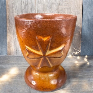May include: A handcrafted, brown ceramic vase with a unique shape. The vase features a wide, open top and a decorative, star-like design carved into the front. The base is rounded, and the overall design is rustic and elegant.