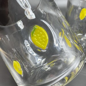 May include: Clear glass pitcher with yellow lemon designs. The pitcher has a wide base and a curved handle. The lemons are painted with yellow and green details. The pitcher is likely used for serving beverages.