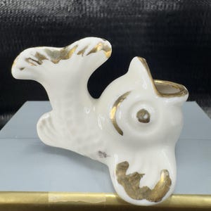May include: A white ceramic fish figurine with gold accents. The fish has an open mouth and a curved tail. The gold detailing is on the fins, tail, and around the mouth. The figurine is on a light blue surface.