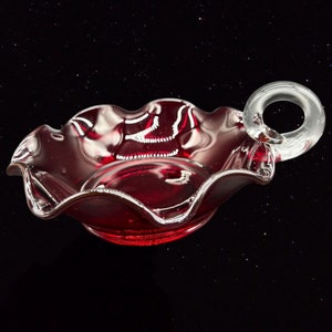 May include: A ruby red glass dish with a clear glass handle. The dish has a scalloped edge and is perfect for serving small snacks or desserts.