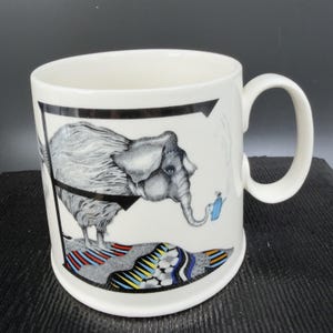 May include: White ceramic mug featuring a whimsical illustration of an elephant with a gray and white textured coat. The elephant is standing on a colorful, patterned surface. A small blue watering can is near the elephant's trunk. The mug has a black border.