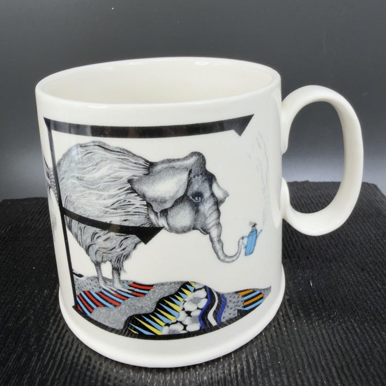 May include: White ceramic mug featuring a whimsical illustration of an elephant with a gray and white textured coat. The elephant is standing on a colorful, patterned surface. A small blue watering can is near the elephant's trunk. The mug has a black border.