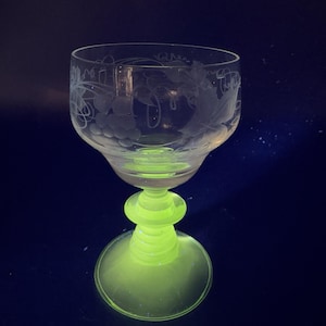 May include: A clear glass goblet with an etched grapevine design and a bright green base. The goblet is on a dark blue background.