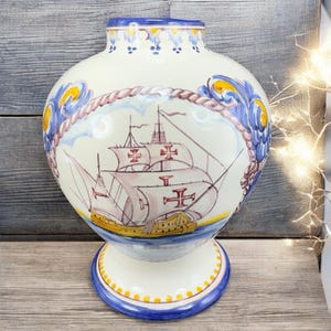 May include: A white ceramic vase with a ship illustration. The vase features blue and yellow decorative accents, including a rope-like border around the ship image. The ship has red cross symbols on its sails. The vase has a pedestal base.