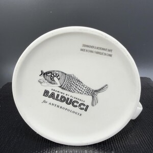 May include: White ceramic mug bottom featuring a black and white fish illustration. The text "DRAWING BY FLORENCE BALDUCCI for ANTHROPOLOGIE" is printed below the fish. The mug is dishwasher and microwave safe.