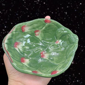 May include: A green ceramic dish with a raised design of leaves and red flowers. The dish is shaped like a leaf and has a slightly rounded bottom.