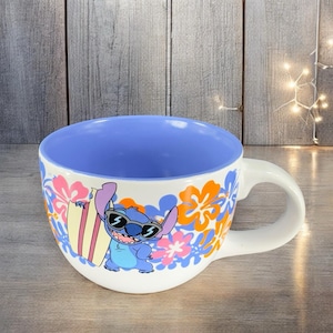 May include: A white ceramic mug with a blue and orange floral design. Stitch, a blue alien character from the Disney movie Lilo & Stitch, is wearing sunglasses and holding a surfboard. The inside of the mug is blue.