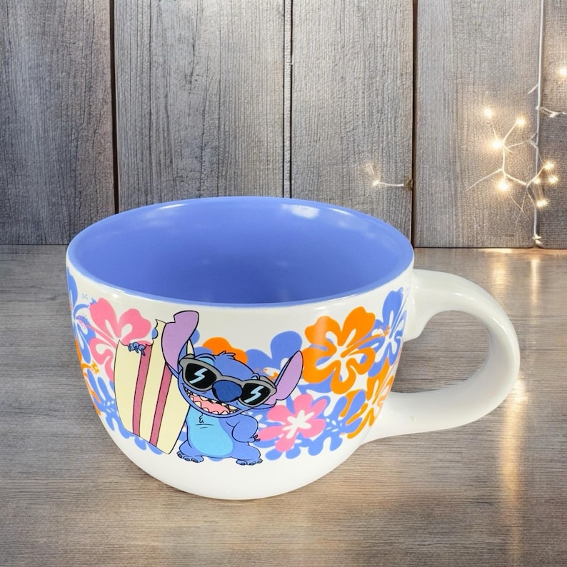 May include: A white ceramic mug with a blue and orange floral design. Stitch, a blue alien character from the Disney movie Lilo & Stitch, is wearing sunglasses and holding a surfboard. The inside of the mug is blue.