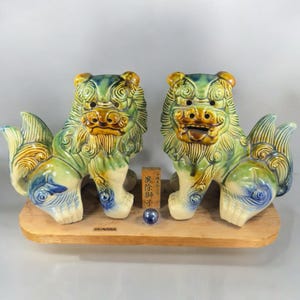May include: A pair of green, yellow, and blue ceramic lion statues with intricate details. The statues are standing on a wooden base with the word "Okinawa" inscribed on it. The statues are traditional Japanese guardian lions, known as Komainu.
