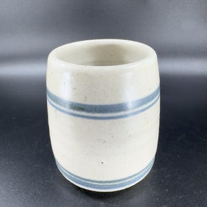 May include: A cylindrical ceramic tumbler with a speckled off-white glaze. Two horizontal blue stripes encircle the body of the tumbler. The interior is a lighter off-white color.