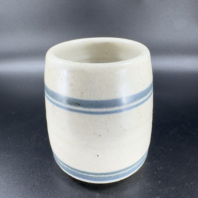 May include: A cylindrical ceramic tumbler with a speckled off-white glaze. Two horizontal blue stripes encircle the body of the tumbler. The interior is a lighter off-white color.