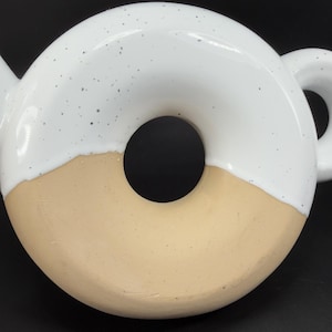 Peut inclure: Une th&eacute;i&egrave;re en c&eacute;ramique en forme de donut. La moiti&eacute; sup&eacute;rieure est blanche avec des d&eacute;tails mouchet&eacute;s, tandis que la moiti&eacute; inf&eacute;rieure est de couleur beige. La th&eacute;i&egrave;re a une anse et un bec, et le design g&eacute;n&eacute;ral est moderne et ludique.