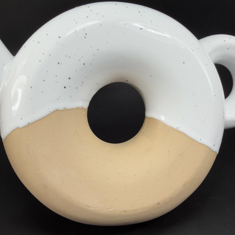 Peut inclure: Une th&eacute;i&egrave;re en c&eacute;ramique en forme de donut. La moiti&eacute; sup&eacute;rieure est blanche avec des d&eacute;tails mouchet&eacute;s, tandis que la moiti&eacute; inf&eacute;rieure est de couleur beige. La th&eacute;i&egrave;re a une anse et un bec, et le design g&eacute;n&eacute;ral est moderne et ludique.