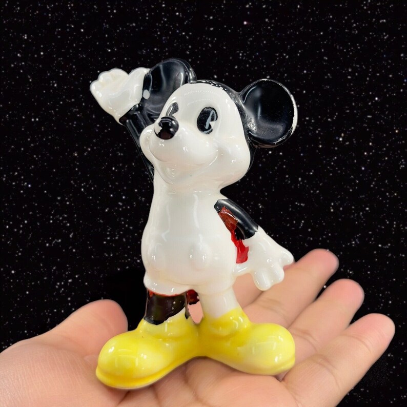 K&ouml;nnte beinhalten: Eine wei&szlig;e Keramikfigur von Mickey Mouse mit schwarzen Ohren und Schuhen. Mickey tr&auml;gt rote Shorts und hat ein schwarz-wei&szlig;es Design auf seinem K&ouml;rper. Er steht mit erhobenem rechten Arm und dem linken Arm an seiner Seite.