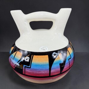 May include: A ceramic vessel featuring a white upper section with two spouts connected by a bridge handle. The lower rounded body displays a vibrant gradient of rainbow colors, including orange, blue, and pink, overlaid with bold black geometric shapes.