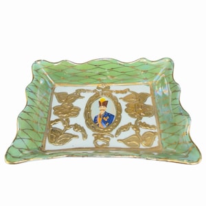 May include: A rectangular, decorative dish with a scalloped edge. The dish features a light green border with a gold lattice pattern. The center has a white background with a portrait of a man in a gold frame, surrounded by gold floral designs.