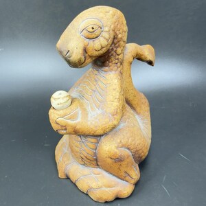 May include: A ceramic sculpture of a stylized creature, possibly a dragon or dinosaur, in shades of brown and tan. The creature holds a small, round object. The sculpture has textured details and a whimsical design, suitable for home decor.