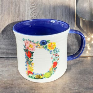 Puede incluir: Taza de cer&aacute;mica blanca con interior y asa azules. La taza presenta un dise&ntilde;o floral en forma de la letra "D". Las flores son de varios colores, incluyendo rosa, amarillo y rojo. La taza tiene una textura acanalada.
