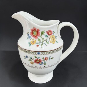 May include: A white ceramic pitcher featuring a colorful floral pattern with red, yellow, and blue flowers. The design includes a decorative band of green leaves and brown scrollwork encircling the middle, with a curved handle and a wide spout.
