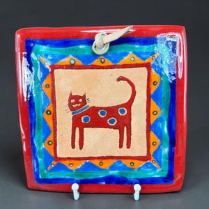 May include: A square, ceramic wall hanging with a red border and a central image of a red cat with blue spots. The cat has a blue collar. The background is tan, and the border has blue, orange, and green accents. A leather loop is attached for hanging.