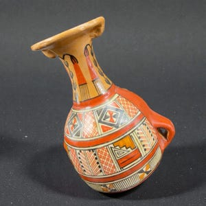 May include: A hand-painted ceramic vase with a curved neck and a handle. The vase features geometric patterns in red, black, and orange against a tan background. The design includes diamond, triangle, and cross motifs, with a flared rim.