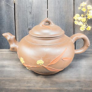 May include: Brown ceramic teapot with a lid, spout, and handle. The teapot is decorated with orange leaf and yellow floral designs. The teapot is sitting on a wooden surface.