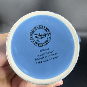 May include: Blue ceramic mug with a Disney Store logo and text that reads "Genuine + Original Disney Store Authentic" and "&copy;Disney Made in Thailand Fabriqu&eacute; en Tha&iuml;lande F784-1014-1-15091".