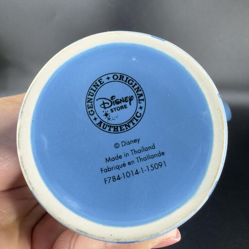 May include: Blue ceramic mug with a Disney Store logo and text that reads "Genuine + Original Disney Store Authentic" and "&copy;Disney Made in Thailand Fabriqu&eacute; en Tha&iuml;lande F784-1014-1-15091".