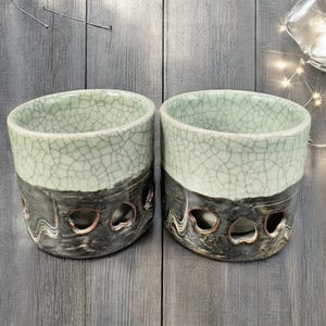 May include: Two ceramic cups with a light green, crackled glaze on the top half and a dark gray, textured bottom half. The bottom half has heart-shaped cutouts. The cups are on a gray wooden surface.