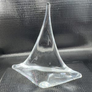 May include: Clear glass sculpture with a triangular shape and a pointed top. The sculpture has a wide base and a smooth, polished surface. The glass is transparent, allowing light to pass through, and it sits on a dark surface.