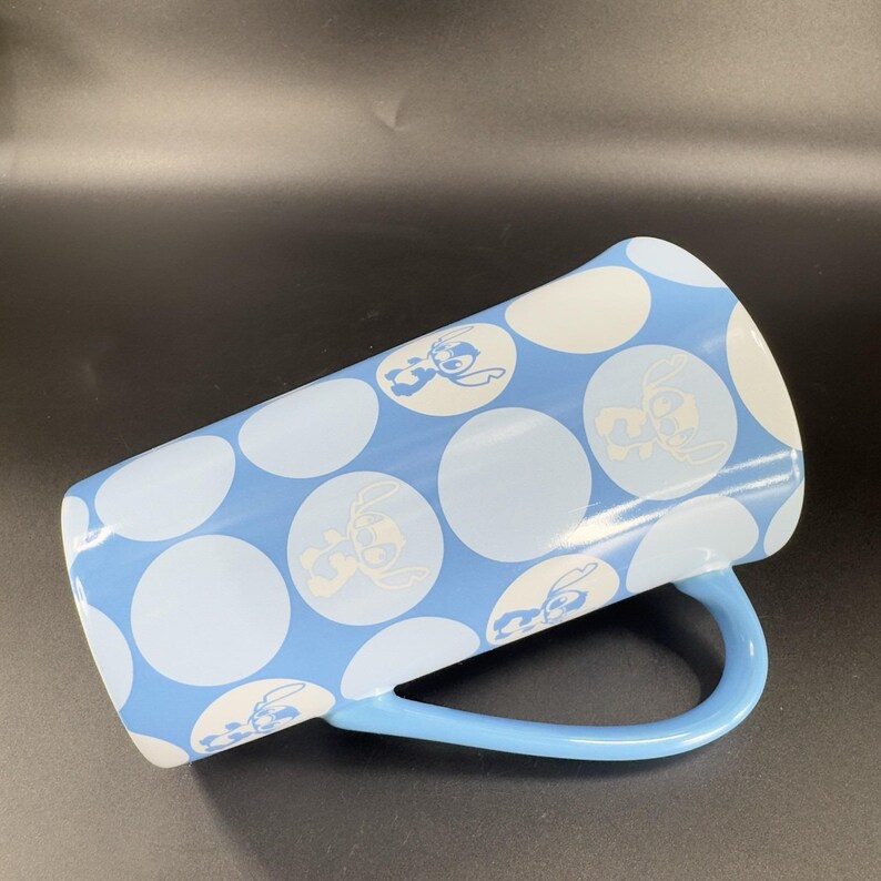 May include: A tall blue ceramic mug with a white polka dot pattern and a light blue handle. The mug features a repeated design of a cartoon character, Stitch, from the Disney movie Lilo & Stitch.