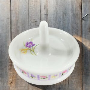 May include: White ceramic ring holder with a floral design. The holder has a raised center post and a base with a purple and white striped pattern.