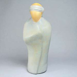 May include: A ceramic sculpture of a figure with a blindfold. The figure is draped in a light blue robe with a yellow face and a white blindfold. The sculpture has a smooth, matte finish and stands on a solid base.