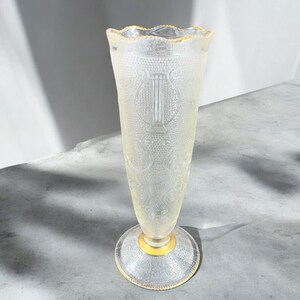 May include: A clear glass vase with a gold rim and intricate etched designs. The vase has a wide base and a narrow neck.