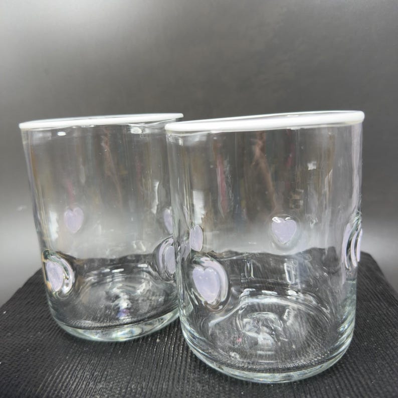 May include: Two clear glass tumblers with a white rim and lavender heart-shaped accents. The glasses are cylindrical and appear to be handmade. The glasses are sitting on a black surface.