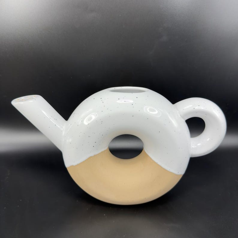 Peut inclure: Une th&eacute;i&egrave;re en c&eacute;ramique en forme de donut. La moiti&eacute; sup&eacute;rieure est blanche avec des mouchetures noires, et la moiti&eacute; inf&eacute;rieure est beige. Elle a un bec verseur et une poign&eacute;e arrondie. La th&eacute;i&egrave;re est un objet d&eacute;coratif.