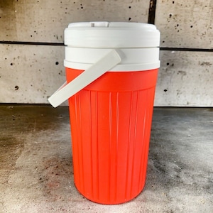 May include: A red and white insulated beverage jug with a white handle. The jug has a ribbed design and a white lid.