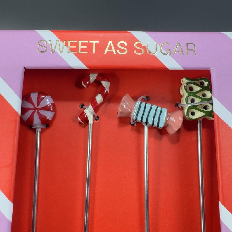 May include: A set of four candy-themed cocktail stirrers. Each stirrer features a different candy design: a red and white swirled lollipop, a red and white candy cane, a blue and pink wrapped candy, and a green and gold candy. The box reads "SWEET AS SUGAR."