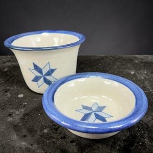 May include: Two ceramic bowls with a white base and blue starburst design. The rims are painted blue. One bowl is a small, shallow dish, and the other is a taller, flared cup.