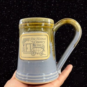 May include: A blue and brown ceramic mug with a gold square logo that reads "Die Heimat Country Inn". The logo features a small house and a wagon with wheels.